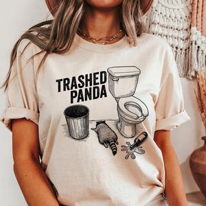 May include: Beige t-shirt with the text "Trashed Panda" in black. The graphic features a raccoon, a toilet, a trash can, and a spilled bottle. The design is a humorous take on a raccoon's antics.