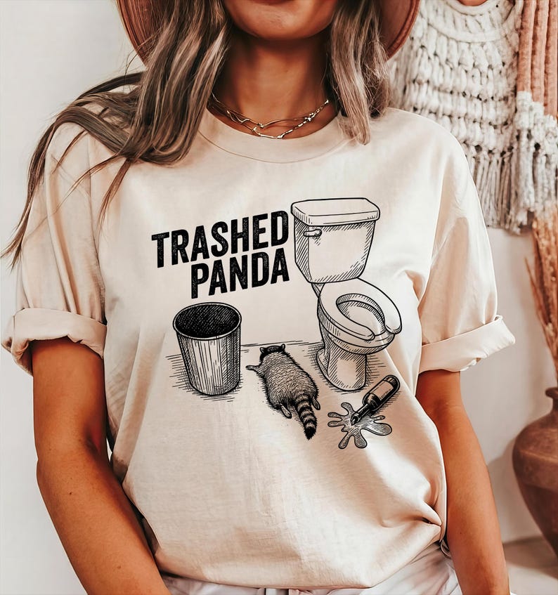 May include: Beige t-shirt with the text "Trashed Panda" in black. The graphic features a raccoon, a toilet, a trash can, and a spilled bottle. The design is a humorous take on a raccoon's antics.