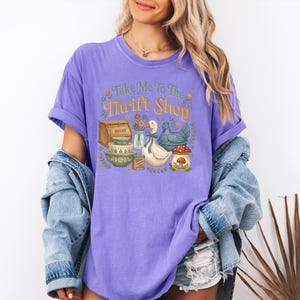 May include: A lilac-colored t-shirt with the phrase "Take Me To The Thrift Shop" in a whimsical font. The graphic features a bread box, bowls, a duck, a chicken, and a mushroom. The model is wearing a denim jacket and shorts.