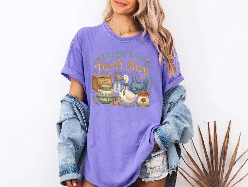 May include: A lilac-colored t-shirt with the phrase "Take Me To The Thrift Shop" in a whimsical font. The graphic features a bread box, bowls, a duck, a chicken, and a mushroom. The model is wearing a denim jacket and shorts.