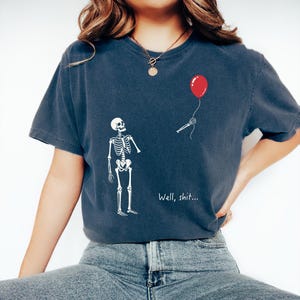 Pode incluir: Uma t-shirt de manga curta azul-escura com um gr&aacute;fico branco de um esqueleto a olhar para um bra&ccedil;o esquel&eacute;tico separado que segura um bal&atilde;o vermelho. O texto "Well, shit..." est&aacute; impresso a branco por baixo do bra&ccedil;o. Gola redonda.