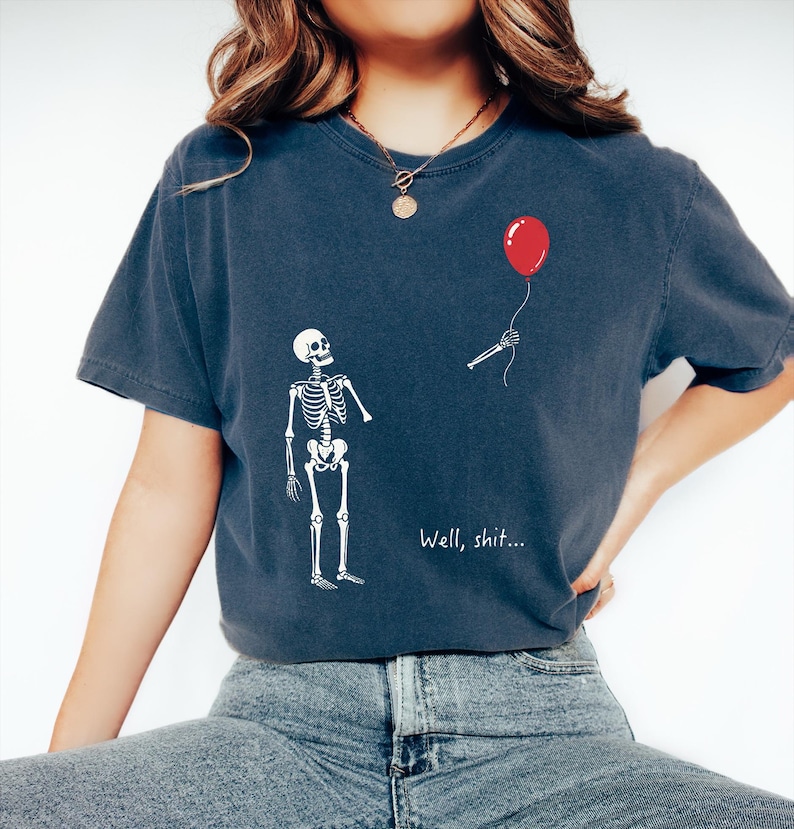 Pode incluir: Uma t-shirt de manga curta azul-escura com um gr&aacute;fico branco de um esqueleto a olhar para um bra&ccedil;o esquel&eacute;tico separado que segura um bal&atilde;o vermelho. O texto "Well, shit..." est&aacute; impresso a branco por baixo do bra&ccedil;o. Gola redonda.