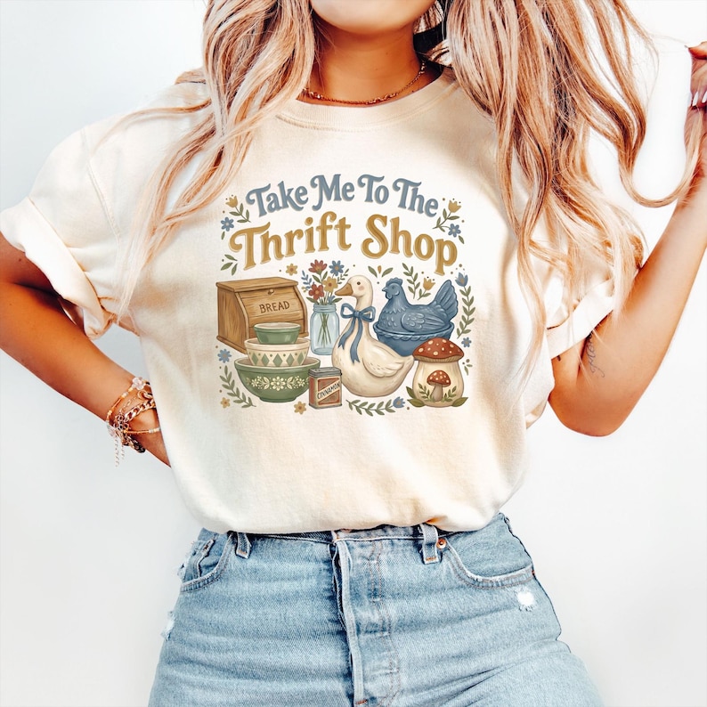 May include: Cream-colored t-shirt with a vintage-style graphic that reads "Take Me To The Thrift Shop." The graphic features a bread box, bowls, a duck, a chicken, and a mushroom.