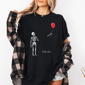 Pode incluir: Uma t-shirt preta com um gr&aacute;fico branco de um esqueleto a olhar para uma m&atilde;o esquel&eacute;tica separada que segura um bal&atilde;o vermelho. O texto "Well, shit..." est&aacute; impresso a branco por baixo do gr&aacute;fico. A t-shirt &eacute; usada com uma camisa de flanela aos quadrados preta e bege.