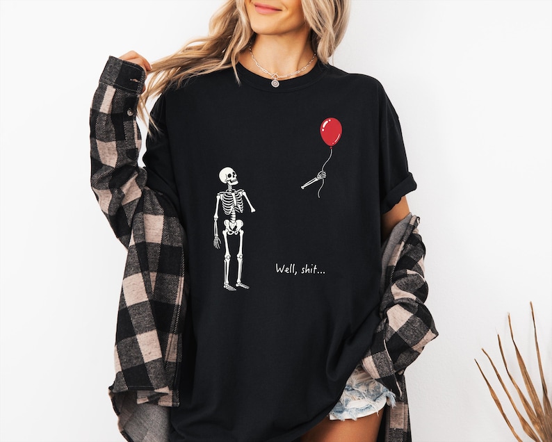 Pode incluir: Uma t-shirt preta com um gr&aacute;fico branco de um esqueleto a olhar para uma m&atilde;o esquel&eacute;tica separada que segura um bal&atilde;o vermelho. O texto "Well, shit..." est&aacute; impresso a branco por baixo do gr&aacute;fico. A t-shirt &eacute; usada com uma camisa de flanela aos quadrados preta e bege.