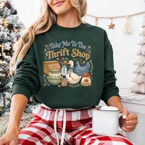 May include: Dark green sweatshirt with the text "Take Me To The Thrift Shop" and a graphic of a duck, chicken, bread box, bowls, and a mushroom. The person is wearing red and white plaid pajama pants and holding a white mug.