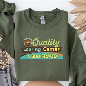 May include: An olive green sweatshirt with the text "Quality Learing Center" in a playful font, with a graphic of a treehouse. Below the text is the phone number 1-800-FRAUD. The sweatshirt is made of a soft material.