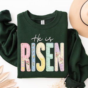 May include: Dark green sweatshirt with the words "He is RISEN" in a colorful patchwork design. The letters are made of various floral and gingham patterns. The text "He is" is written in a white script font.
