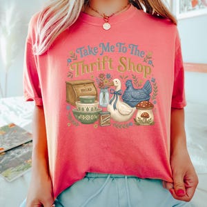 May include: Coral-colored t-shirt with the text "Take Me To The Thrift Shop" in blue and yellow. The graphic includes a bread box, bowls, a duck, a chicken, and a mushroom. Floral accents surround the design.