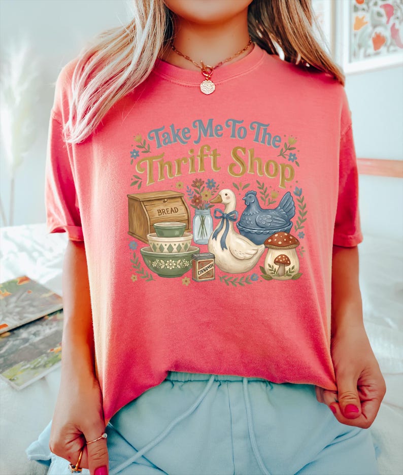 May include: Coral-colored t-shirt with the text "Take Me To The Thrift Shop" in blue and yellow. The graphic includes a bread box, bowls, a duck, a chicken, and a mushroom. Floral accents surround the design.