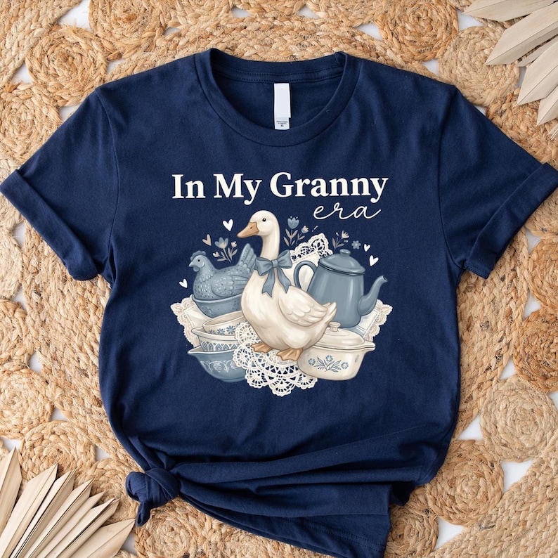 An illustration of a white duck wearing a blue ribbon, surrounded by a blue hen figurine, vintage blue and white bowls, a blue kettle, and lace doilies with the text "In My Granny Era" at the top.