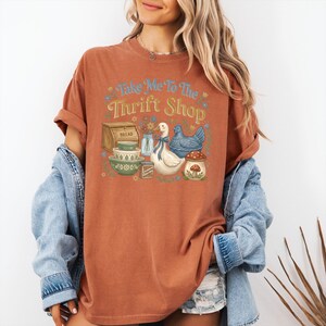 May include: Rust-colored t-shirt with the text "Take Me To The Thrift Shop" and a graphic of a bread box, bowls, a duck, a chicken, and a mushroom. The shirt has short sleeves and a crew neck.