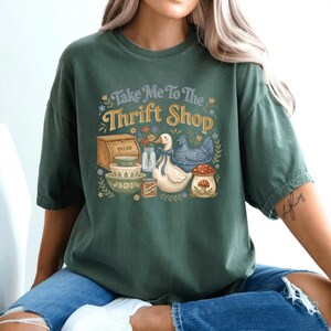 May include: Dark green t-shirt with the text "Take Me To The Thrift Shop" in a vintage style. The graphic includes illustrations of a bread box, bowls, a duck, a chicken, and a mushroom.