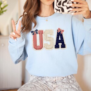 May include: A pale blue sweatshirt featuring the letters "USA" in a decorative appliqu&eacute;. The letters are adorned with red and white stripes, navy blue polka dots, and lace edging. A casual, comfortable garment.