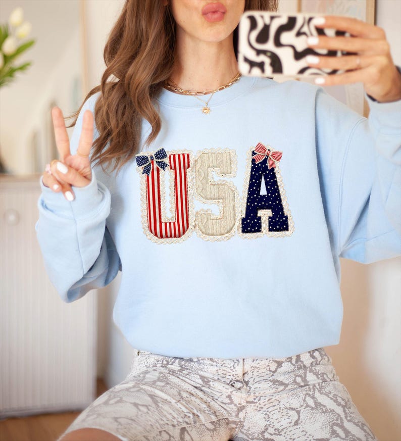 May include: A pale blue sweatshirt featuring the letters "USA" in a decorative appliqu&eacute;. The letters are adorned with red and white stripes, navy blue polka dots, and lace edging. A casual, comfortable garment.