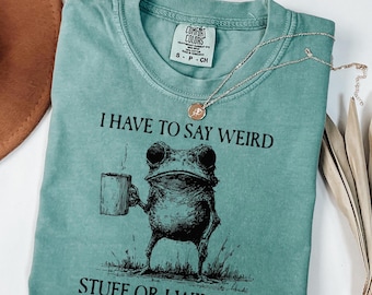 May include: A teal Comfort Colors t-shirt with the text "I HAVE TO SAY WEIRD STUFF OR I WILL DIE" and a frog illustration holding a mug. A silver necklace with a pendant is draped across the shirt.