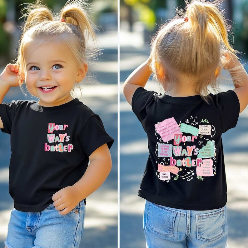 A young girl with blonde pigtails smiles while wearing a black short-sleeved shirt. The front features a colorful text design that reads Your Ways Better. The back of the shirt displays a collage of colorful notes with various faith-based messages and botanical illustrations surrounding the central text.