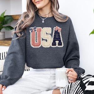May include: Dark grey sweatshirt featuring the word "USA" in a patriotic design. The letters are decorated with red and white stripes, navy blue polka dots, and cream-coloured lace trim. A bow accents the top of the letters.