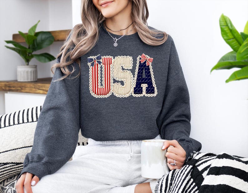 May include: Dark grey sweatshirt featuring the word "USA" in a patriotic design. The letters are decorated with red and white stripes, navy blue polka dots, and cream-coloured lace trim. A bow accents the top of the letters.