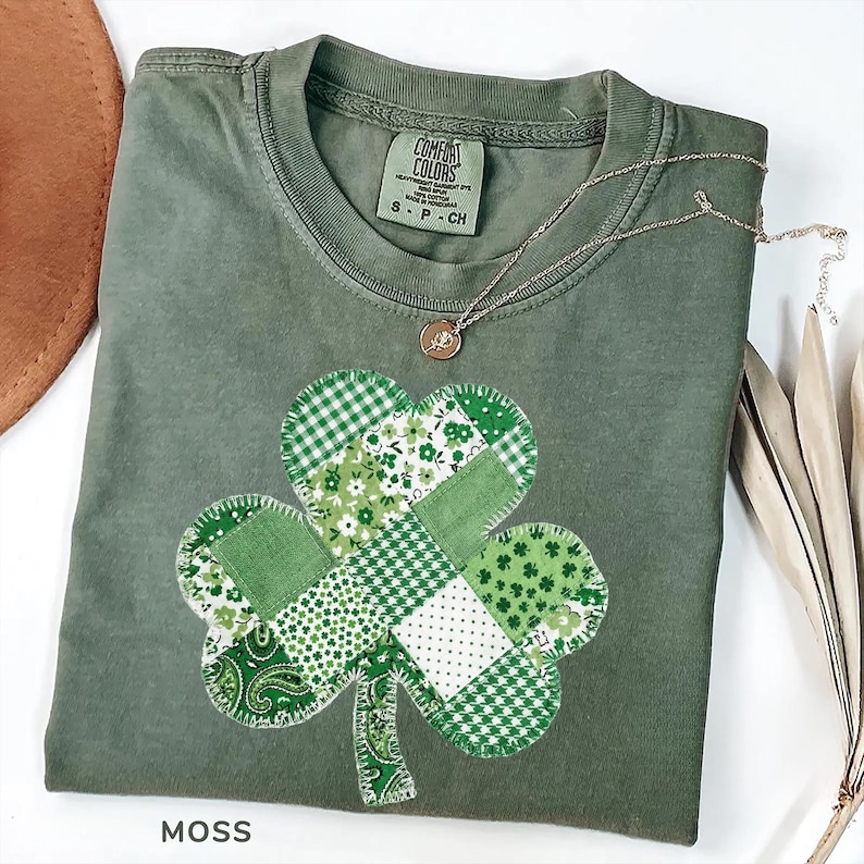 A moss green t-shirt with a patchwork shamrock design. The shamrock is made of various green patterned fabrics. A gold necklace with a small pendant rests on the shirt. The shirt is labeled "Comfort Colors" and the word "MOSS" is at the bottom.