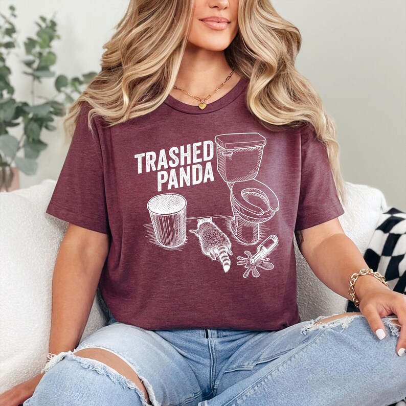 May include: Burgundy t-shirt with the text "Trashed Panda" and a white graphic of a raccoon, toilet, and trash can. The shirt is a short-sleeved crew neck.
