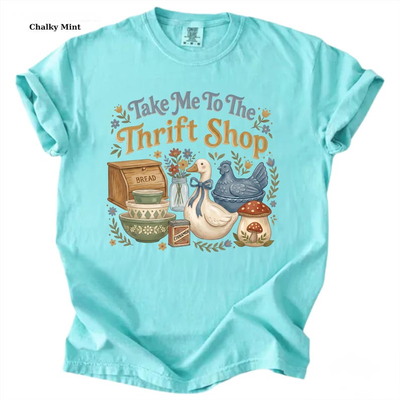 May include: A light blue t-shirt with the text "Take Me To The Thrift Shop" in a whimsical font. The graphic includes a wooden bread box, bowls, a goose, a chicken, and a mushroom. The shirt is made of a soft material.