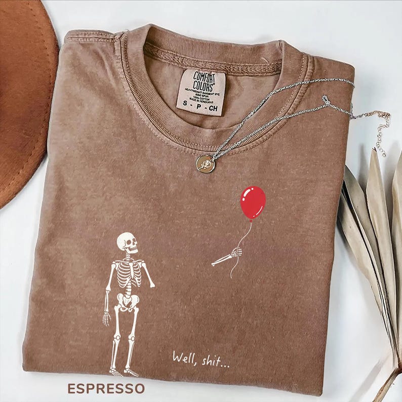 Pode incluir: Uma t-shirt castanha de gola redonda com etiqueta Comfort Colors, apresentando um gr&aacute;fico branco de um esqueleto e um bal&atilde;o vermelho. O texto "Well, shit..." est&aacute; impresso a branco por baixo do esqueleto. Um colar prateado repousa na gola.