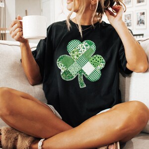 May include: Black t-shirt featuring a large, patchwork shamrock design in various shades of green and white. The shamrock is composed of different patterns, including gingham, floral, and paisley. The person is holding a white mug.