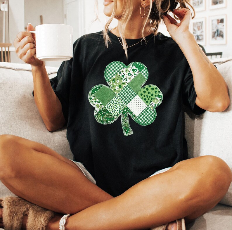 May include: Black t-shirt featuring a large, patchwork shamrock design in various shades of green and white. The shamrock is composed of different patterns, including gingham, floral, and paisley. The person is holding a white mug.