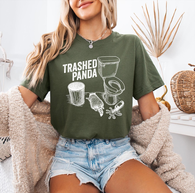 May include: Olive green t-shirt with the text "Trashed Panda" above a graphic of a toilet, trash can, raccoon, and spilled items. The design is in white. The shirt is a casual, unisex style.