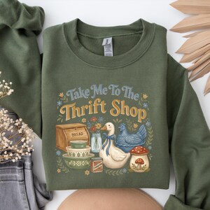 May include: Olive green sweatshirt with the phrase "Take Me To The Thrift Shop" in blue and gold lettering. The graphic includes a duck, bread box, bowls, and a mushroom. The sweatshirt has long sleeves and a crew neck.