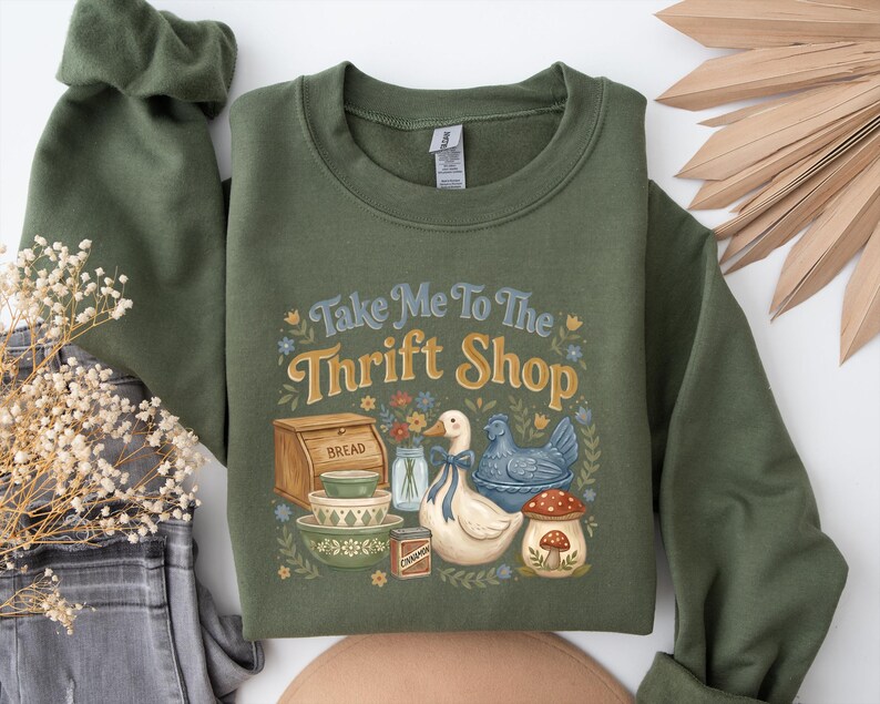 May include: Olive green sweatshirt with the phrase "Take Me To The Thrift Shop" in blue and gold lettering. The graphic includes a duck, bread box, bowls, and a mushroom. The sweatshirt has long sleeves and a crew neck.