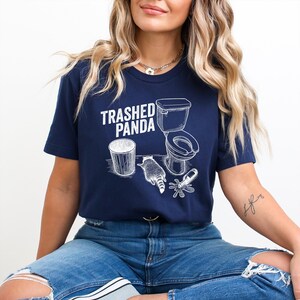 May include: Navy blue t-shirt with a white graphic of a raccoon, toilet, and trash can, with the text "Trashed Panda". The design is a humorous take on a raccoon's behavior. The shirt is made of a soft material.