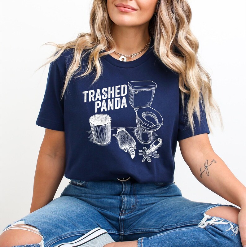 May include: Navy blue t-shirt with a white graphic of a raccoon, toilet, and trash can, with the text "Trashed Panda". The design is a humorous take on a raccoon's behavior. The shirt is made of a soft material.