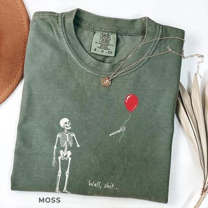 Pode incluir: Uma t-shirt de gola redonda verde musgo com um gr&aacute;fico branco de um esqueleto de p&eacute; a olhar para um bal&atilde;o vermelho flutuante segurado por uma m&atilde;o esquel&eacute;tica desencarnada. O texto "Well, shit..." est&aacute; impresso a branco sob o gr&aacute;fico.