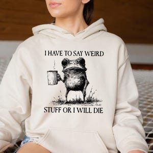 May include: Cream-colored hoodie with a front pocket and drawstring hood. The graphic features a frog holding a mug with steam rising, and the text "I HAVE TO SAY WEIRD STUFF OR I WILL DIE" in black.