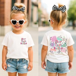 A young girl with blonde pigtails smiles while wearing a black short-sleeved shirt. The front features a colorful text design that reads Your Ways Better. The back of the shirt displays a collage of colorful notes with various faith-based messages and botanical illustrations surrounding the central text.