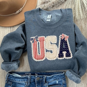 May include: A grey sweatshirt featuring the word "USA" in a decorative applique. The letters are adorned with red and white stripes, blue polka dots, and bows. A brown hat and blue jeans are also visible.