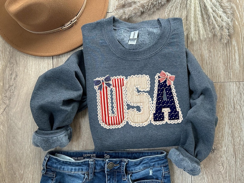 May include: A grey sweatshirt featuring the word "USA" in a decorative applique. The letters are adorned with red and white stripes, blue polka dots, and bows. A brown hat and blue jeans are also visible.