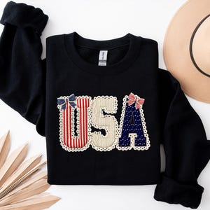May include: Black sweatshirt featuring the word "USA". The letters are decorated with red and white stripes, blue polka dots, and a beige textured fabric, with small bows and a scalloped lace trim.