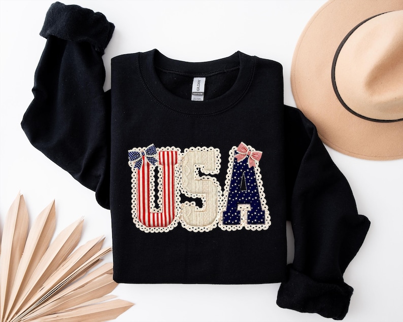May include: Black sweatshirt featuring the word "USA". The letters are decorated with red and white stripes, blue polka dots, and a beige textured fabric, with small bows and a scalloped lace trim.