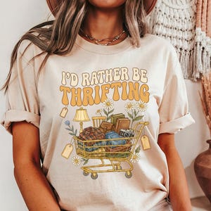 Op de afbeelding: Beige t-shirt met de tekst "I'd Rather Be Thrifting" in oranje en geel. De afbeelding toont een winkelwagentje vol vintage items zoals boeken, een lamp en kleding, met madeliefjes.
