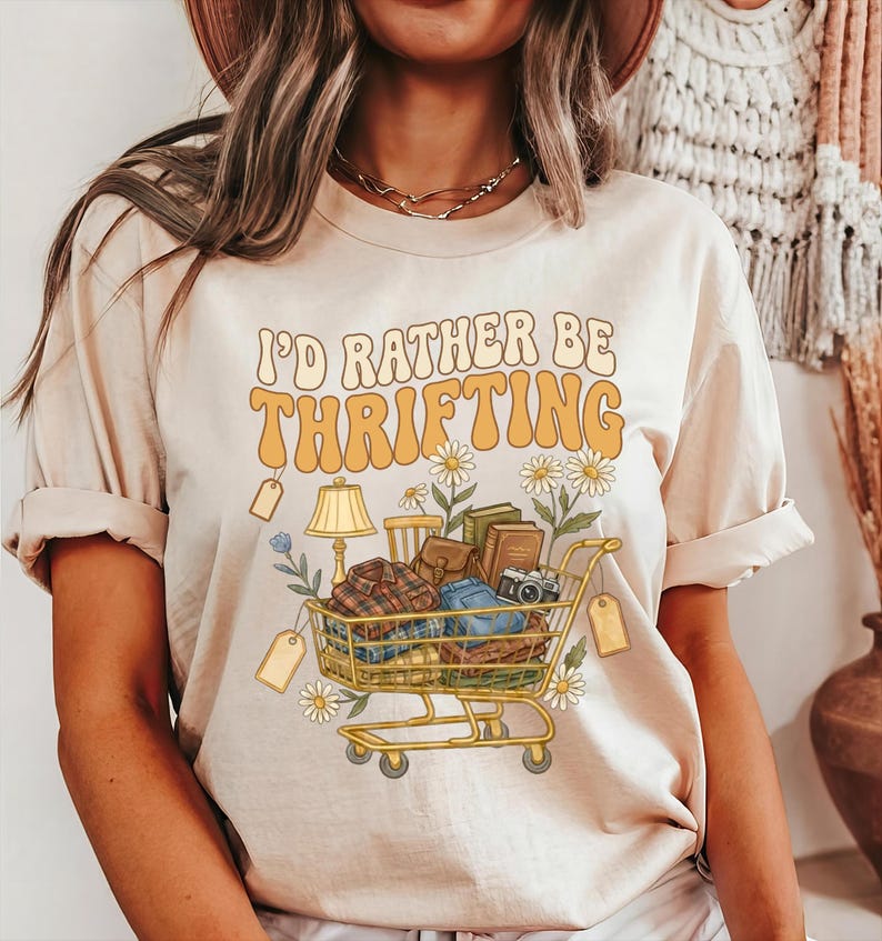 Op de afbeelding: Beige t-shirt met de tekst "I'd Rather Be Thrifting" in oranje en geel. De afbeelding toont een winkelwagentje vol vintage items zoals boeken, een lamp en kleding, met madeliefjes.