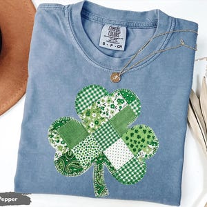 May include: A light blue Comfort Colors t-shirt with a patchwork shamrock design in various shades of green. A gold necklace with a pendant rests on the shirt. The shirt is size S-P-CH.