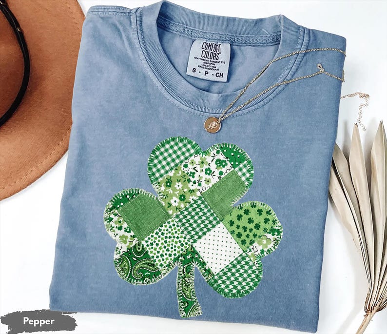 May include: A light blue Comfort Colors t-shirt with a patchwork shamrock design in various shades of green. A gold necklace with a pendant rests on the shirt. The shirt is size S-P-CH.