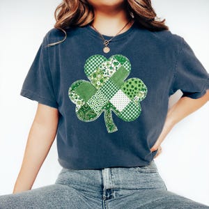 May include: A navy blue t-shirt featuring a patchwork shamrock design in various shades of green and white. The shamrock is centered on the front of the shirt, with a mix of patterns including gingham, floral, and paisley. The shirt has short sleeves and a relaxed fit.