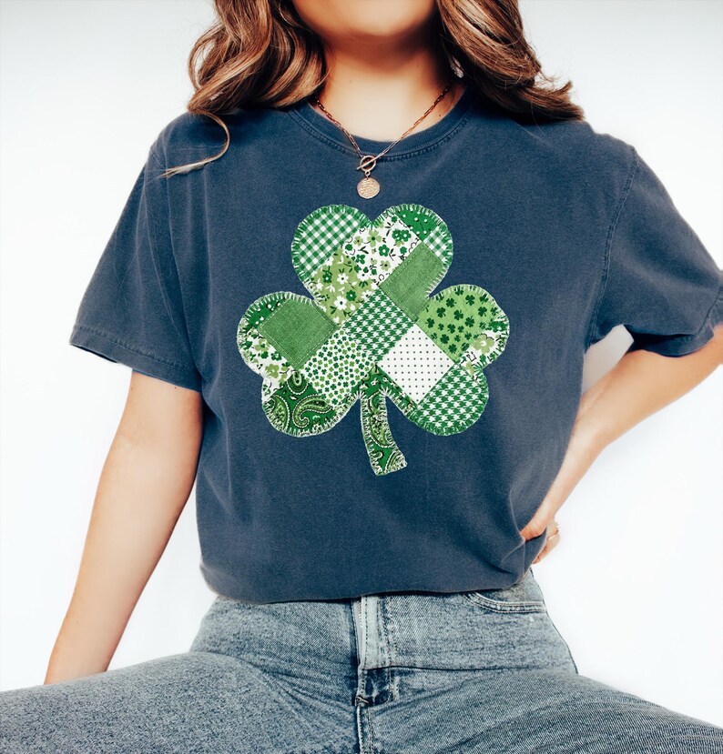 May include: A navy blue t-shirt featuring a patchwork shamrock design in various shades of green and white. The shamrock is centered on the front of the shirt, with a mix of patterns including gingham, floral, and paisley. The shirt has short sleeves and a relaxed fit.