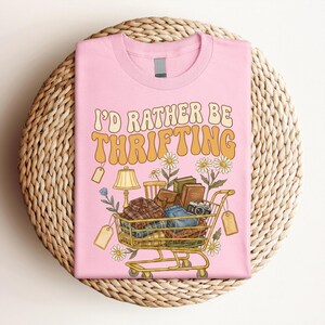 Op de afbeelding: Roze T-shirt met de tekst "I'D RATHER BE THRIFTING" in oranje en geel. De afbeelding toont een winkelwagentje vol kleding, boeken en een camera, samen met madeliefjes en prijskaartjes.