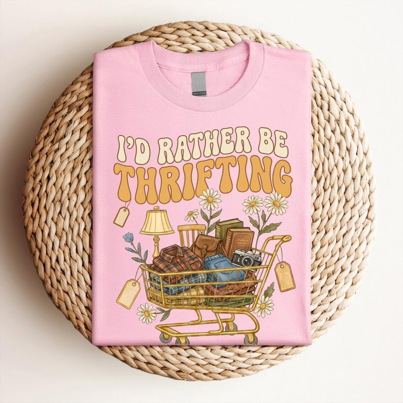 Op de afbeelding: Roze T-shirt met de tekst "I'D RATHER BE THRIFTING" in oranje en geel. De afbeelding toont een winkelwagentje vol kleding, boeken en een camera, samen met madeliefjes en prijskaartjes.