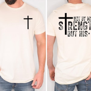 May include: Cream-colored t-shirt with a black cross on the front and back. The back of the shirt features the text "NOT BY MY STRENGTH BUT HIS" in a distressed font. The shirt is a short-sleeved crew neck.